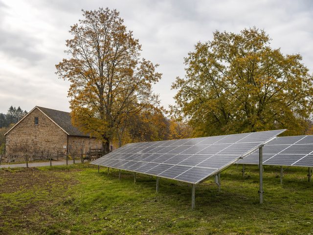 Rangées de panneaux au sol sur pieux vissés dans une prairie, derrière une clôture, bâtiment agricole en fond